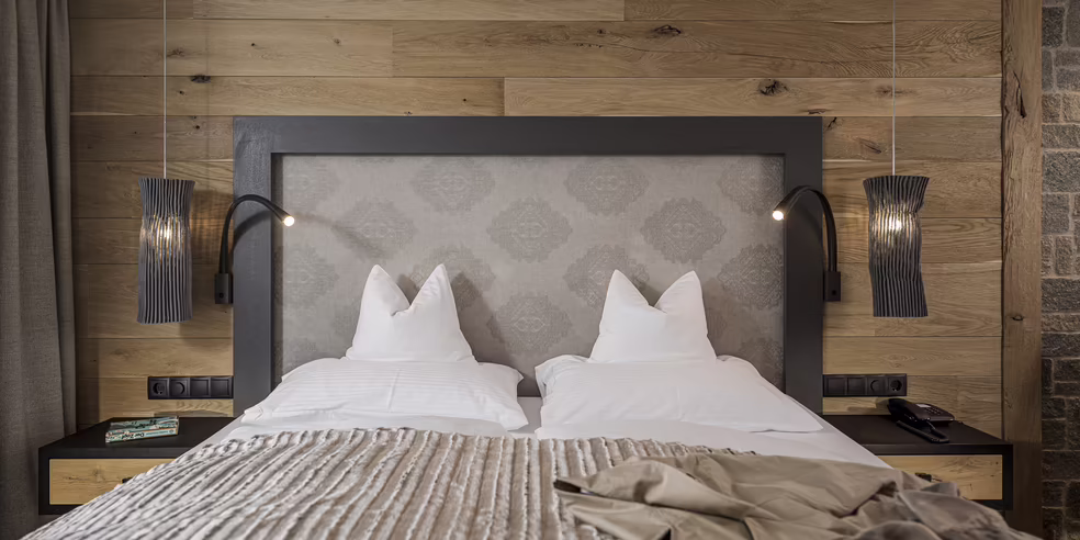 Entspannt schlafen in der Wohlfühlsuite Maya im Hotel Platzlhof Ein modernes Schlafzimmer mit einem Doppelbett, Holzvertäfelung an der Wand und stilvollen Nachttischlampen.