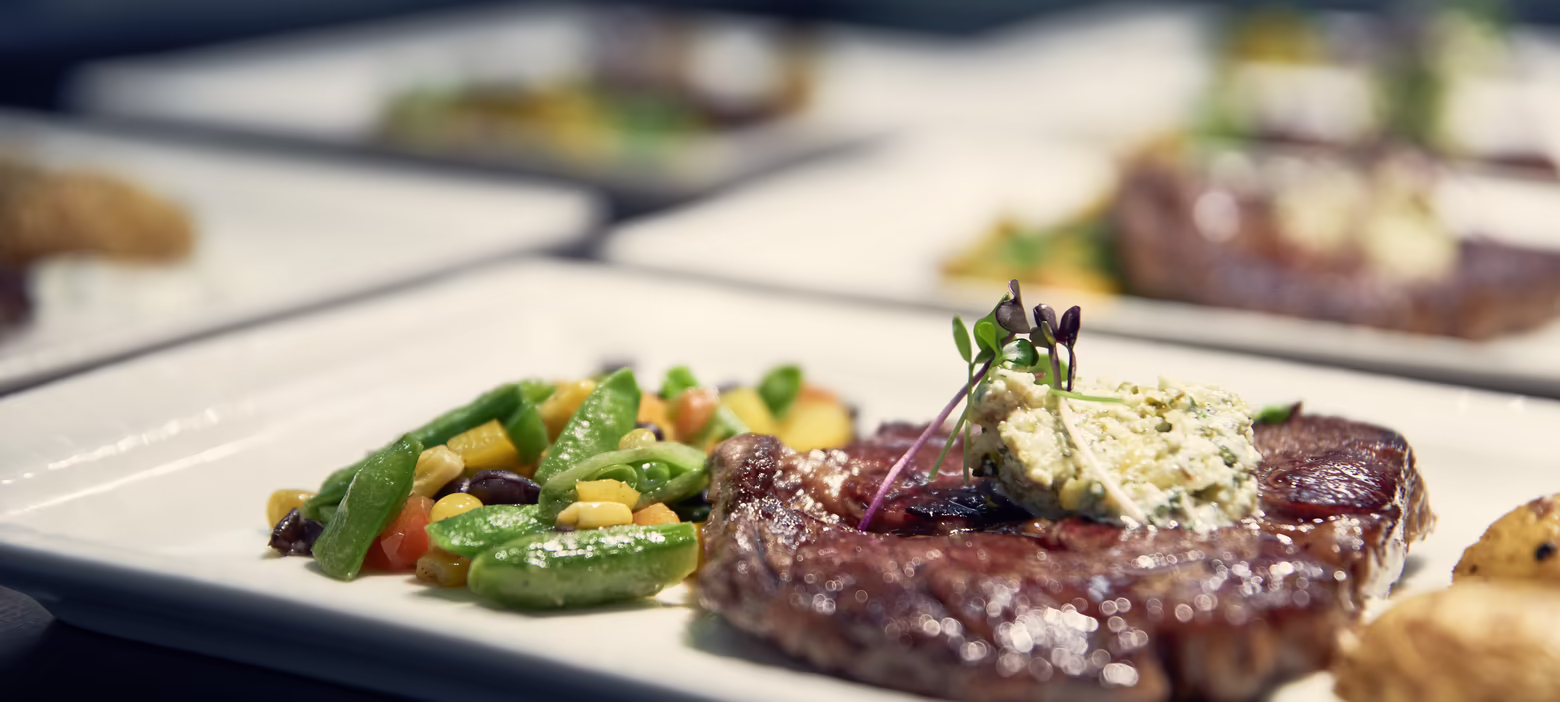 Essen im Hotel Platzlhof Ein Teller mit saftigem Steak, garniert mit Kräuterbutter, und einer Beilage aus buntem Gemüse. Weitere Teller im Hintergrund.